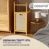 A bedroom scene featuring a wooden shelf unit with a laundry basket, a bedside table with a lamp, and a bed with gray bedding. Produktbild-Vorschau