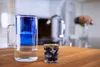 A glass water pitcher and a glass of water with berries and ice cubes are on a wooden countertop in a kitchen. Produktbild-Vorschau