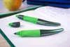 Two green pens are placed on an open notebook with blue ink writing. An apple and a blue pencil case are visible in the background. Produktbild-Vorschau