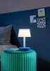 A small white lamp sits on a green textured table next to a bed. Produktbild-Vorschau
