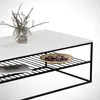 A close-up view of a modern coffee table with a white top and black metal frame, featuring a decorative arrangement of branches in a glass vase and a small bowl of potpourri on the lower shelf. Produktbild-Vorschau