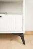 A close-up of a white cabinet with a fluted drawer front and a black angled leg. Produktbild-Vorschau