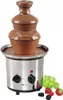 A chocolate fountain with a stainless steel base and a ceramic top, surrounded by grapes and berries. Podgląd zdjęcia produktu