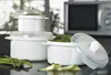 A set of white microwave containers with clear lids, some filled with spinach, are arranged on a dark countertop. Asparagus and zucchini peels are visible in the foreground. A microwave is visible in the background. Produktbild-Vorschau