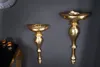 Two ornate gold wall consoles are mounted on a dark gray wall. The console on the left holds a stack of gold rings. The console on the right holds a small dish with some decorative items. To the left, a black table with gold trim holds three decorative can Produktbild-Vorschau
