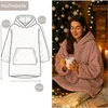 A woman wearing a pink oversized hoodie blanket, holding a mug, in a cozy setting with a Christmas tree and lights. A technical drawing of the hoodie blanket is shown on the left with measurements. Produktbild-Vorschau