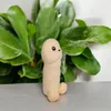 A plush penis toy sits on a gray surface in front of a large green plant. Náhled obrázku produktu