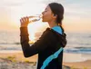 A woman drinks water from a bottle on a beach at sunset. Náhľad obrázku produktu