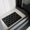 A close-up view of the interior of a fireplace, showing a black metal grate on a white tiled hearth, with the corner of a glass door visible. Produktbild-Vorschau