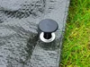 A close-up view of a black plastic grommet with a silver metal ring on a green tarp, with grass in the background. Produktbild-Vorschau