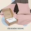 An open cardboard box with a white folded textile inside, placed on a bed with pink bedding. Produktbild-Vorschau