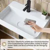 A hand is cleaning a white sink with a gray cloth. A black faucet and a small potted plant are visible. Anteprima dell’immagine prodotto