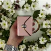 A hand holding a pink mug shaped like a handbag, surrounded by white flowers. Produktbild-Vorschau