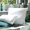 Two pillows are arranged on a bed with a book and glasses in the foreground, near a window with plants and decorative items. Produktbild-Vorschau