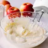 A close-up of a hand mixer beating ingredients in a glass bowl, with apples, eggs, and flour in the background. Produktbild-Vorschau