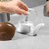 A hand is shown pressing down on the pump of a white snail-shaped soap dispenser, with a brown snail shell object in the foreground. Voir les images du produit