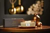 A WoodWick candle and its packaging are displayed on a wooden table, with decorative golden objects in the background. Podgląd zdjęcia produktu
