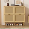 A light wood shoe cabinet with four rattan doors and wooden legs, displayed in a room with shoes underneath. Podgląd zdjęcia produktu