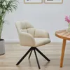 A cream upholstered swivel chair with black metal legs, placed on a wooden floor next to a potted plant and a round wooden table with a vase of pink flowers. Produktbild-Vorschau