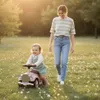 A woman is walking next to a child on a ride-on toy car in a grassy park with flowers. Produktbild-Vorschau