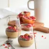 Two glasses of dessert with pomegranate seeds and almonds on a white table, with pomegranates and a mug in the background. Produktbild-Vorschau