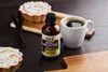A bottle of vanilla extract is placed on a dark wooden surface next to a cup of coffee and two tarts. Náhľad obrázku produktu