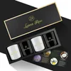 A gift set of four scented candles in a black box with a gold trim and gold lettering. A small booklet with images of lavender, lemons, and coconuts is placed in front of the candles. Produktbild-Vorschau