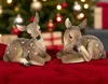 Two fawn figurines are lying down on a red surface, with a blurred Christmas tree and gifts in the background. Produktbild-Vorschau