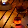 A small, orange-glowing speaker sits on a phone on a wooden table next to a glass of amber liquid. Podgląd zdjęcia produktu