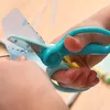 A close-up shot of a child's hands using teal-colored training scissors to cut a piece of paper with a blue design. Voir les images du produit