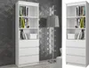 Two white bookshelves are shown side-by-side against a textured gray wall. The bookshelf on the left is more fully visible, displaying books and storage boxes on its shelves, and drawers at the bottom. A floor lamp is positioned between the two bookshelves Produktbild-Vorschau