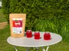 A product bag and three small jars of jelly are placed on a white table outdoors, with a green hedge in the background. Produktbild-Vorschau