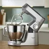 A silver stand mixer with a mixing bowl and attachment is shown on a wooden countertop in a kitchen. Voir les images du produit