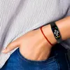 A person's wrist wearing a smart band and a red string bracelet, with part of a striped shirt and jeans visible. Voir les images du produit