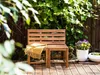 A wooden garden bench with a yellow blanket and potted flowers on a wooden deck, surrounded by greenery. Produktbild-Vorschau