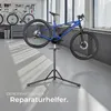 A blue electric bicycle is mounted on a black repair stand in a workshop or garage setting. Tools are visible on a pegboard in the background. Náhled obrázku produktu