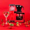 A red Moccacoffee machine is displayed with strawberries on a red background. Náhled obrázku produktu