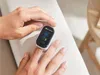 A person's hand is using a pulse oximeter on their finger, which is placed on a textured light-colored surface. Produktbild-Vorschau