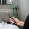 A person holding a black electric toothbrush over a white sink in a bathroom. Anteprima dell’immagine prodotto