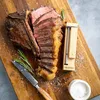 A cooked and sliced steak is presented on a wooden board with a meat thermometer, a knife, rosemary sprigs, and salt. Náhled obrázku produktu
