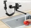 A black kitchen faucet with a spray hose is mounted on a white wall above a stainless steel sink. A hand is shown washing a red tomato under the running water. Náhled obrázku produktu