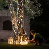 A person is lighting lanterns under a tree decorated with string lights. Podgląd zdjęcia produktu