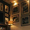 A collection of framed pictures on a wall, with a floor lamp in the foreground. Náhľad obrázku produktu