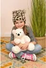 A young girl is sitting cross-legged on a rug, holding a white teddy bear. She is wearing a leopard-print hat and a striped shirt. A tall plant in a basket is to her right. Náhľad obrázku produktu