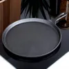 A black cast iron crepe pan is shown on a black reflective surface, with a wooden object and a plant in the background. Voir les images du produit