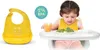 A baby is sitting in a high chair eating broccoli, with a bib on. A yellow bib is shown separately on the left. Produktbild-Vorschau