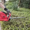 A person wearing a black and white dotted glove is using a red electric hedge trimmer to trim a green hedge. Produktbild-Vorschau
