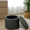 A dark gray storage ottoman with its lid open, placed on a wooden floor next to a chest of drawers and a potted plant in a woven basket. Produktbild-Vorschau
