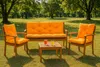 A wooden garden bench and two chairs with orange cushions are set up on a grassy lawn in a park-like setting with trees in the background. Produktbild-Vorschau