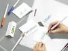 A person's hands are shown writing names on cards with a calligraphy pen. Several pens and cards with names written on them are scattered on a table. Voir les images du produit